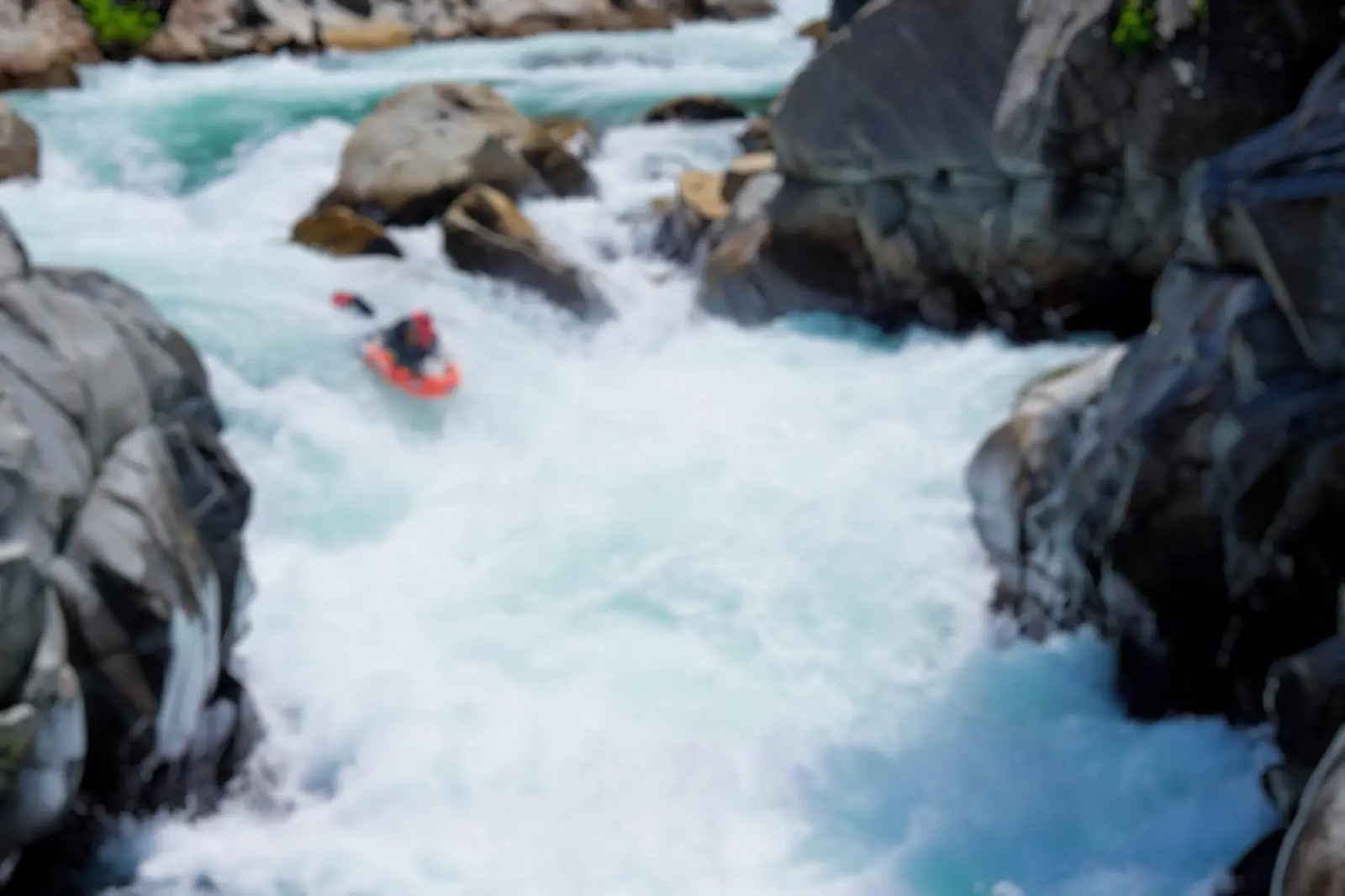 A riverboarder on a red board running a turquoise rapid between dark boulders